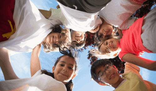 Un alegre grupo de niños tomados de las manos en un círculo, capturado desde abajo con un cielo brillante de fondo.